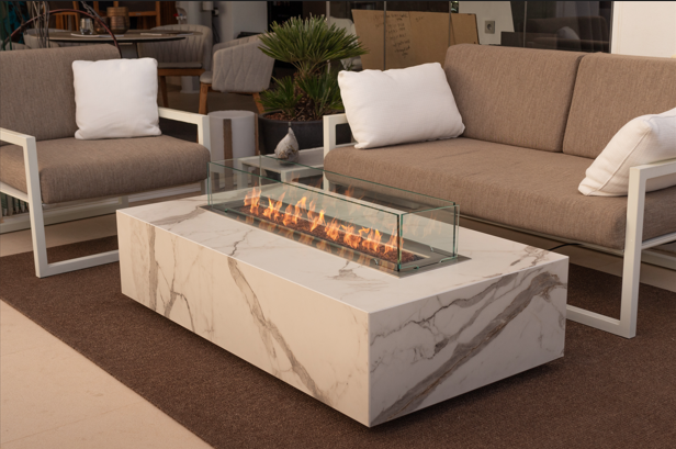 Outdoor seating area with beige cushioned sofas around a rectangular marble fire table featuring a glass-enclosed linear flame.