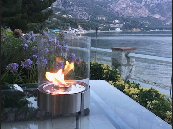 Small cylindrical tabletop fire lantern with a glass enclosure burning a bright flame on a patio table, overlooking coastal water, mountains, and garden flowers at dusk.