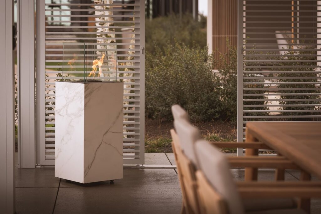 Outdoor patio area with a tall marble pedestal fire feature enclosed in glass, positioned near sliding louvered panels, with wooden dining furniture and greenery in the background.