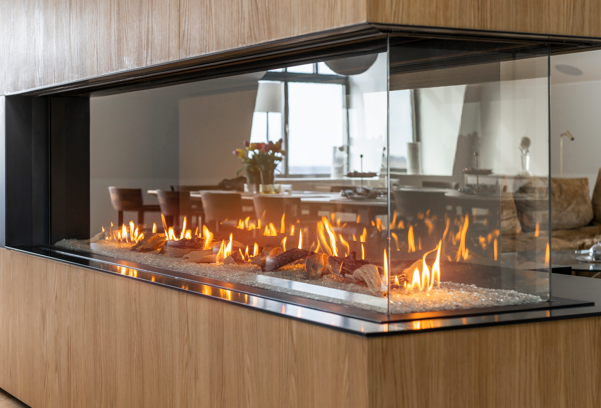 Modern double-sided glass fireplace with visible flames, set in a wood-paneled structure, showing a dining area with chairs and a table in the background.