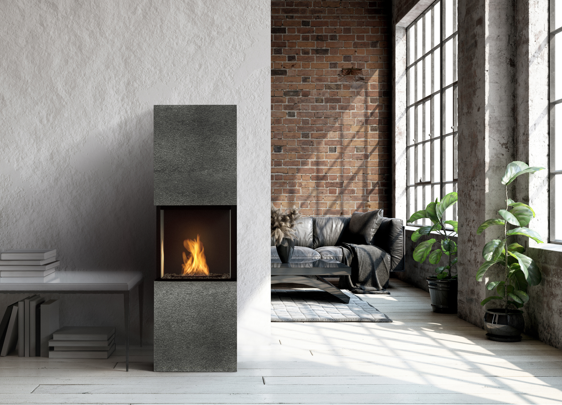 Loft-style interior with large industrial windows, a tall modern fireplace with a single flame, a white textured wall, a brick accent wall, a dark sofa, and several potted plants in natural light.